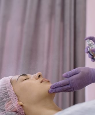 Anonymous cosmetician in protective gloves applying moisturizing spray on face of client lying on couch with closed eyes during beauty procedure